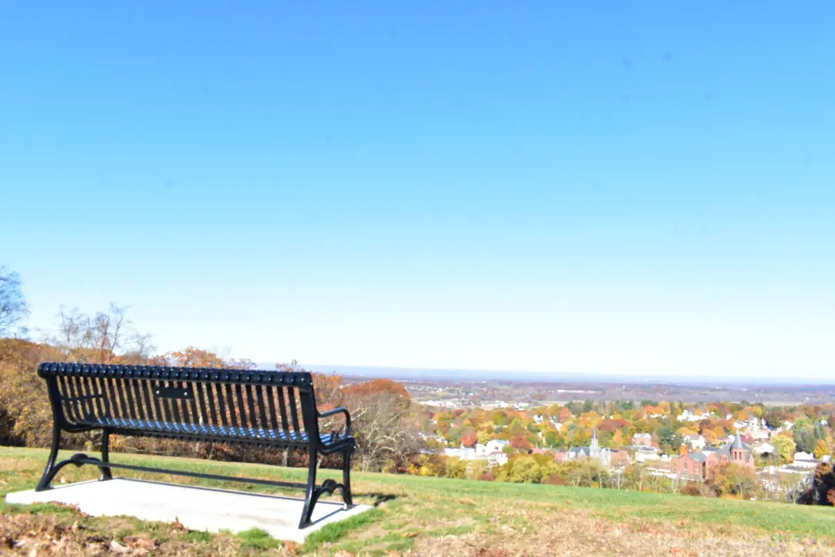 A black metal park bench sits alone on a grassy hill overlooking a colorful autumn town under a clear blue sky. The overlaid…