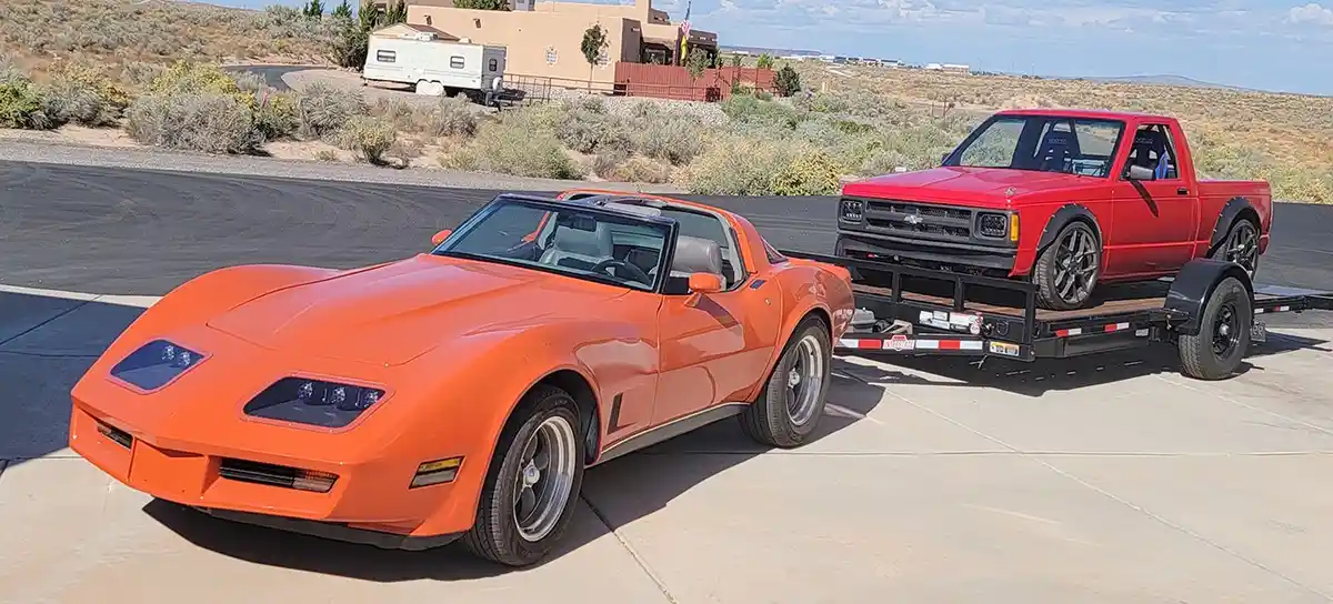 An orange classic Chevrolet Corvette sports car is parked on pavement, towing a flatbed trailer that carries a red pickup…
