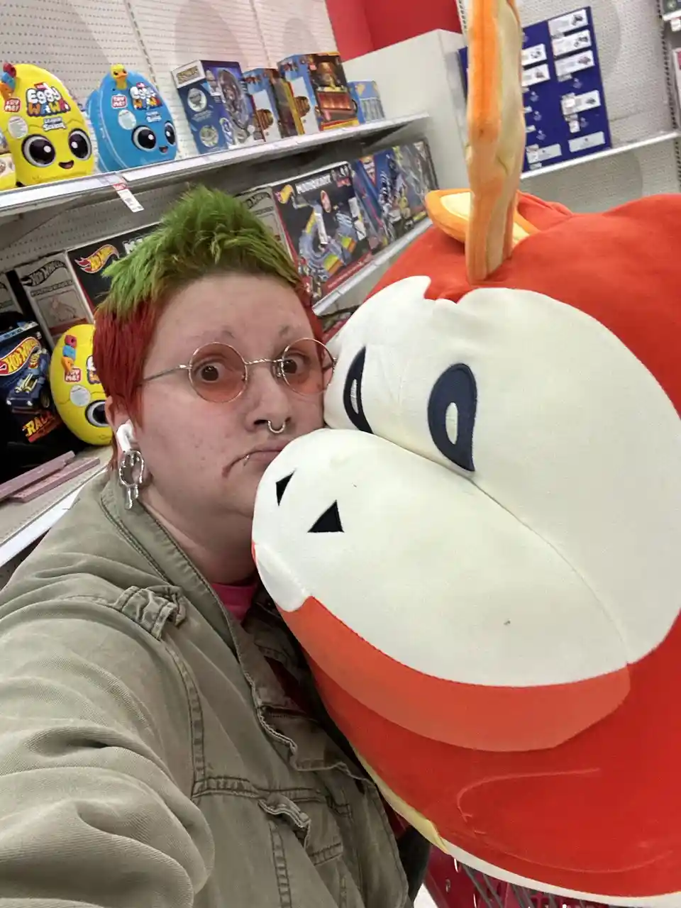 A person with green and red hair and round glasses poses in a toy aisle, cheek-to-cheek with a large red and white plush toy…
