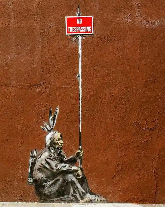 A stencil graffiti artwork depicts a Native American figure sitting on the ground, holding a long pole that extends upward…