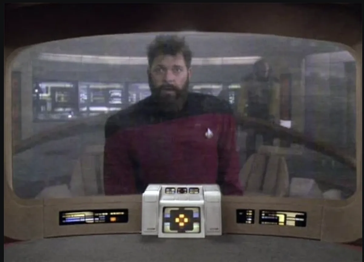 A man with a beard wearing a red Starfleet uniform sits at a spaceship's command console, looking surprised. The console has…