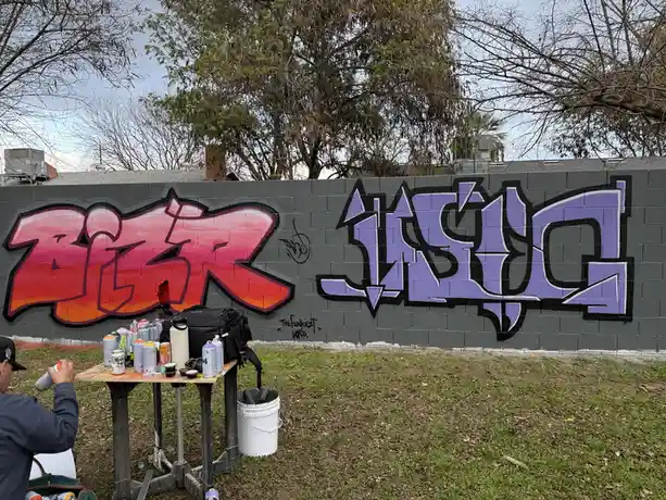 A person is spray painting graffiti on a gray brick wall outdoors, with a table full of spray cans and art supplies nearby…