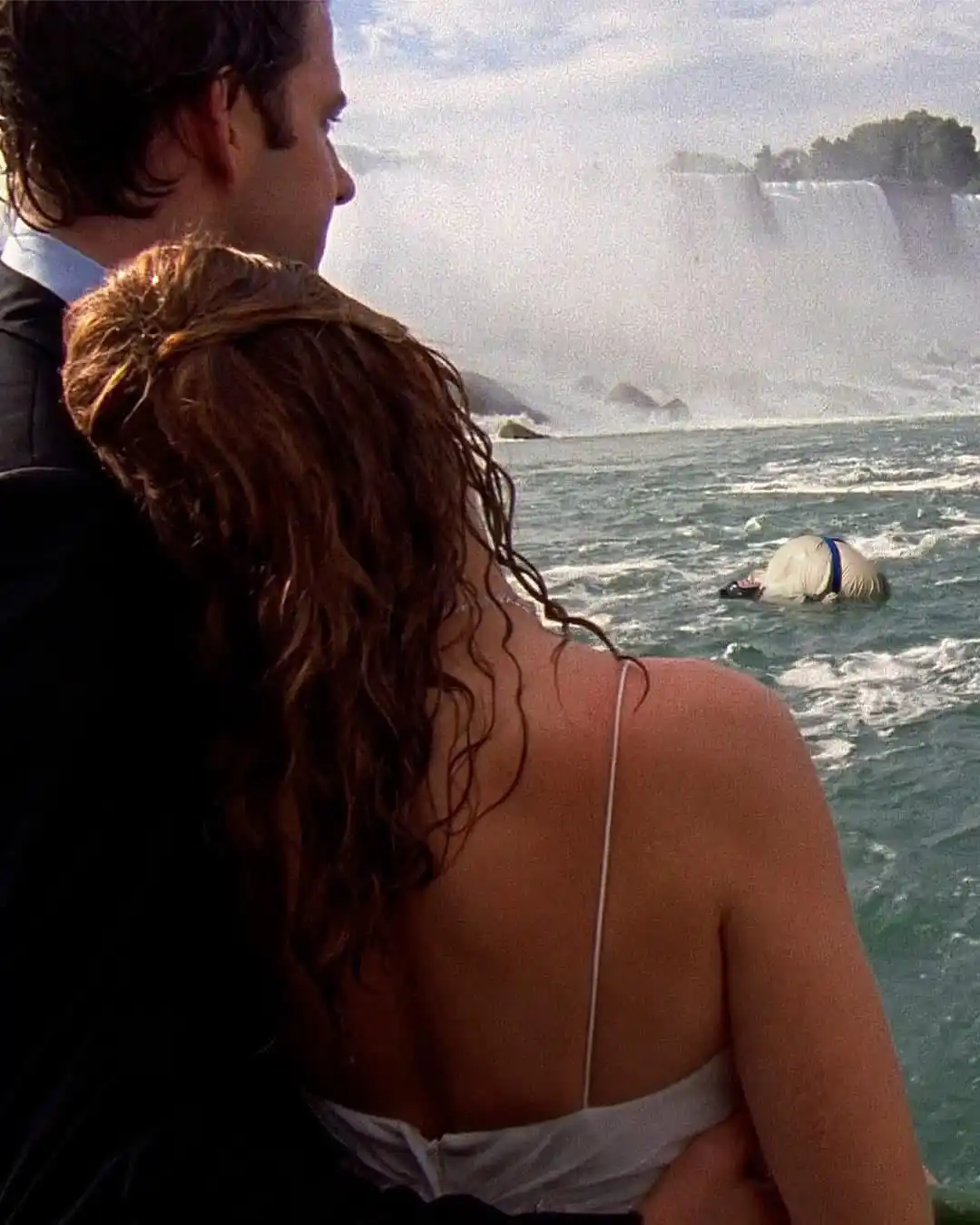 A man and woman in formal attire stand close together, looking out at a large waterfall. In the turbulent water below them…