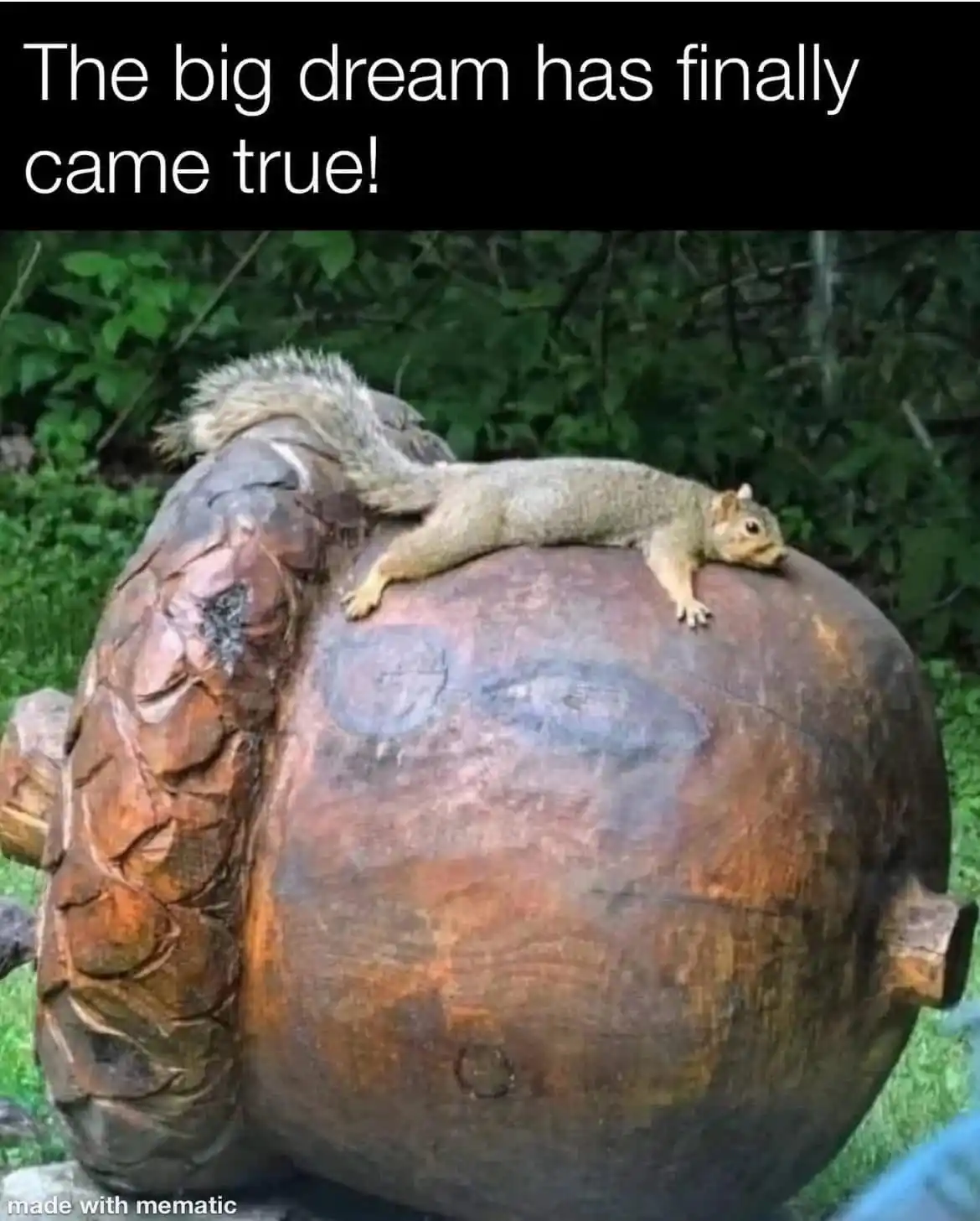 A squirrel is lying stretched out on top of a large, carved wooden sculpture of an acorn, appearing relaxed and content. The…
