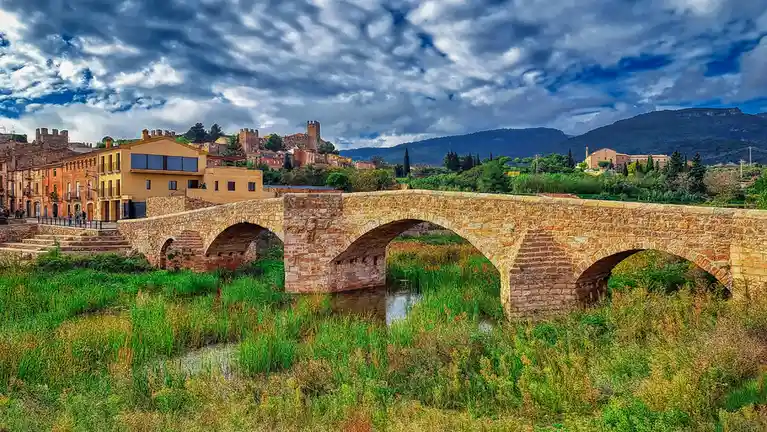 A scenic, heavily saturated photo of a historic stone arch bridge spanning a grassy river, with a European-style town and…