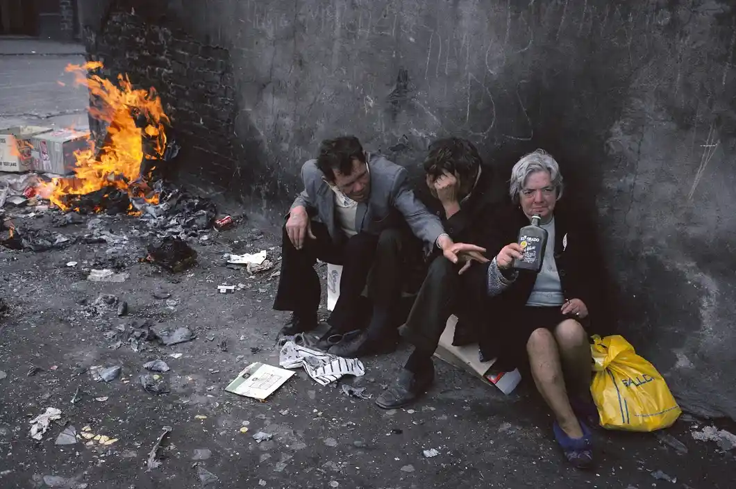 Three people sit against a grimy wall next to a burning pile of trash. The woman on the right holds a bottle of liquor while…