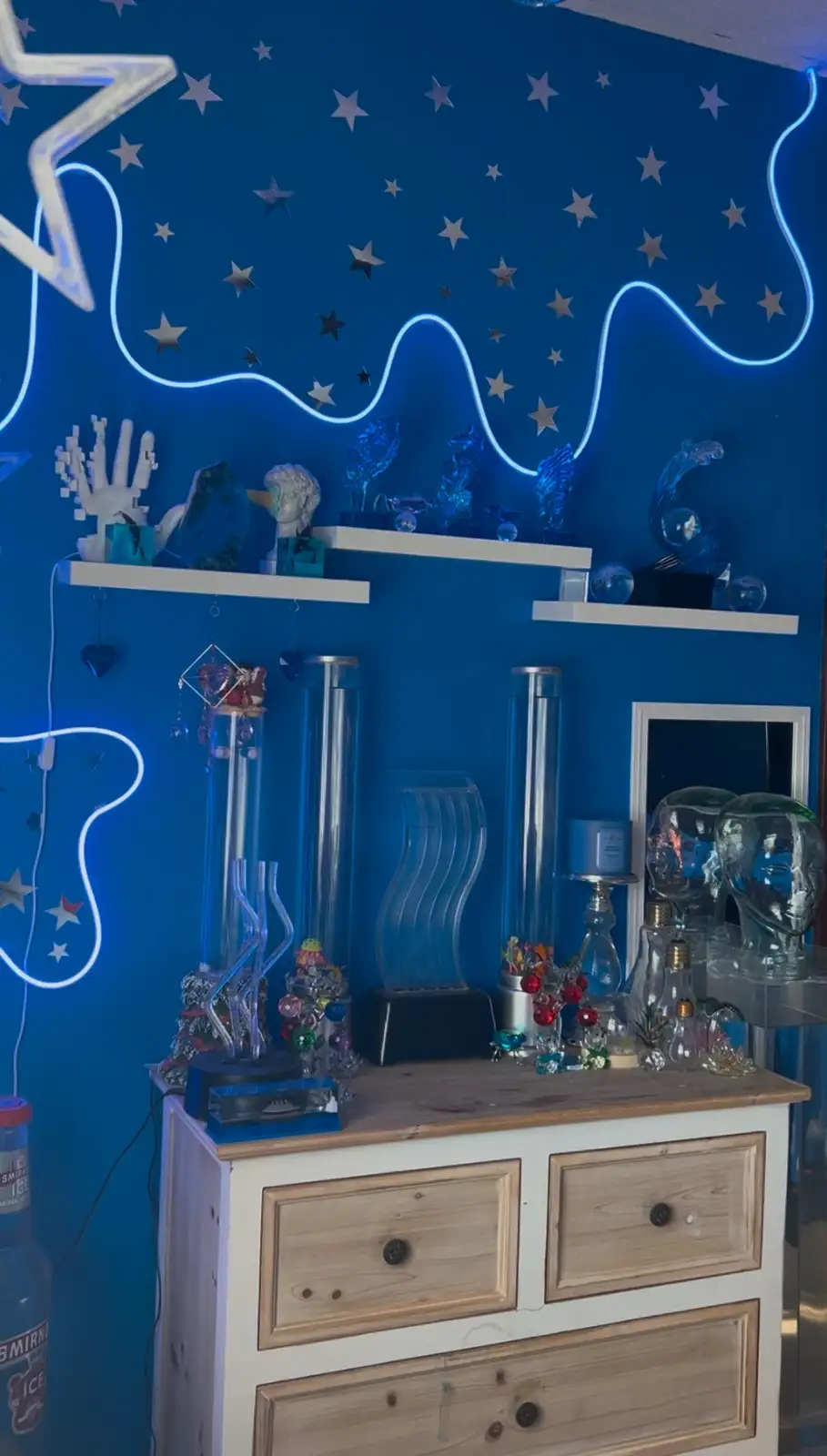 A room with a blue wall covered in star decals and glowing neon lights shaped like a guitar and a wave. White shelves hold…