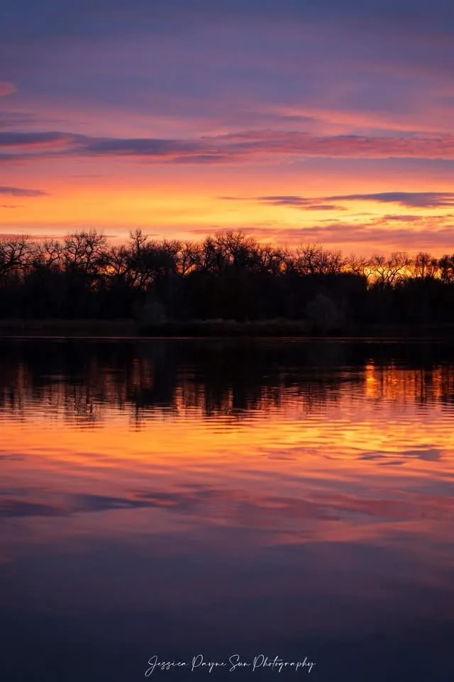 A serene sunset photograph showing vibrant orange, pink, and purple clouds reflected in calm water, with dark silhouettes of…
