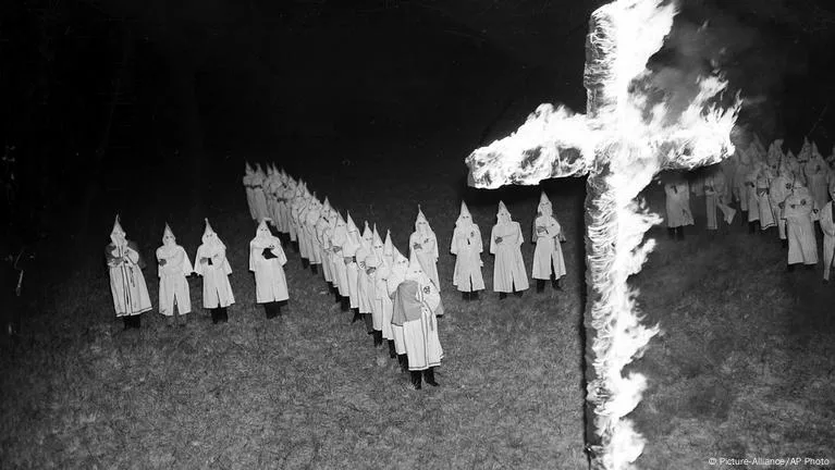 A black-and-white historical photograph shows a group of people in white robes and pointed hoods standing in formation…