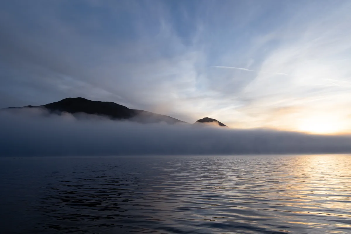 A serene landscape photo shows a calm body of water reflecting a sunrise or sunset, with misty clouds partially obscuring…