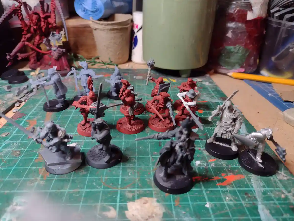 A collection of unpainted and partially painted fantasy miniatures on a green grid mat, with some figures painted red and…
