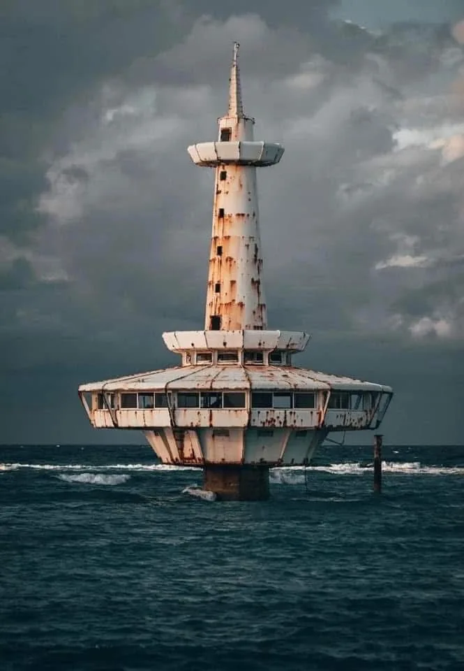 A rusted, white, multi-level structure resembling a lighthouse or offshore platform stands partially submerged in dark…