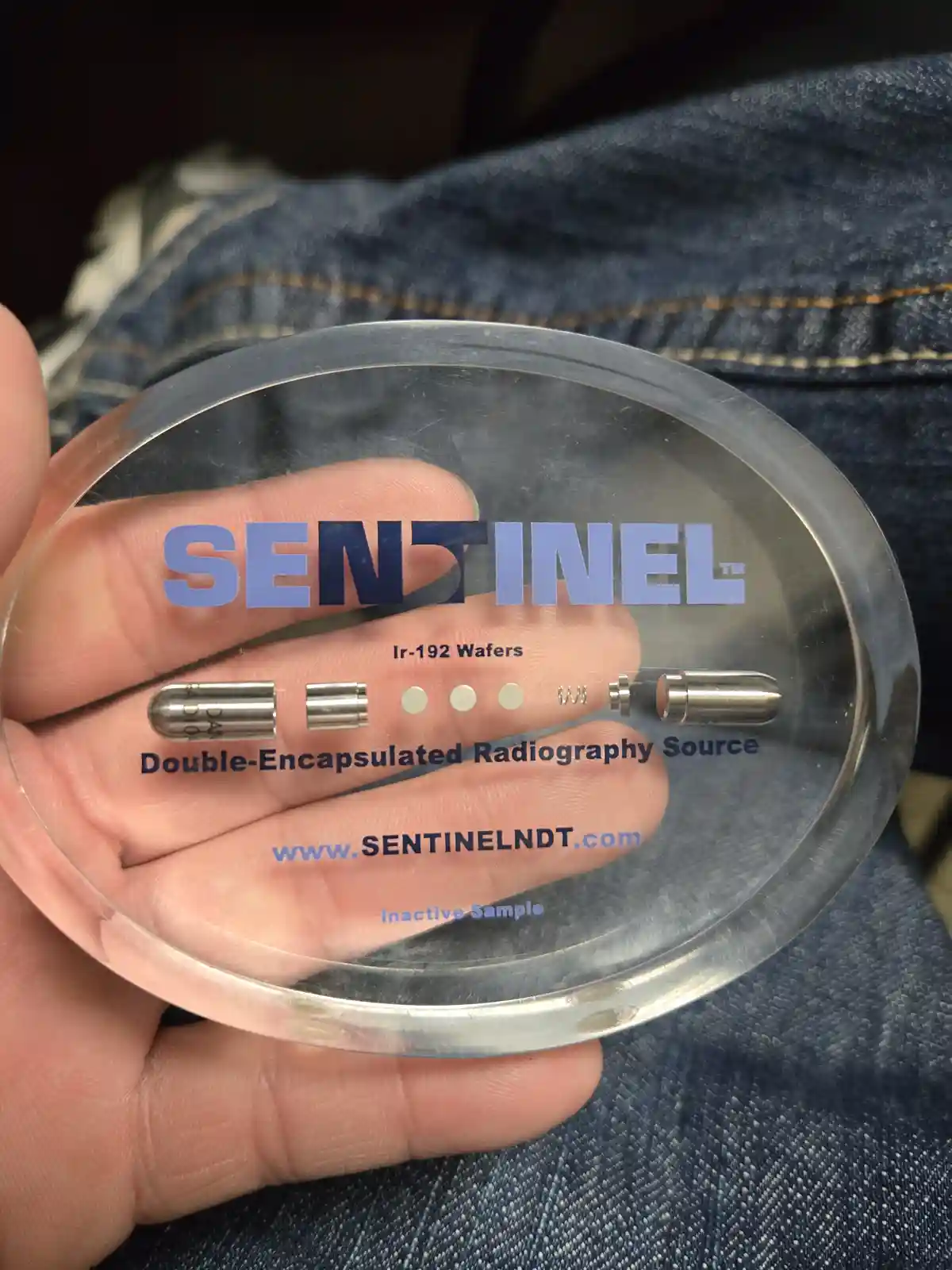 A hand holds a clear, circular plastic case containing small metallic components labeled as 'SENTINEL™ Ir-192 Wafers' and…