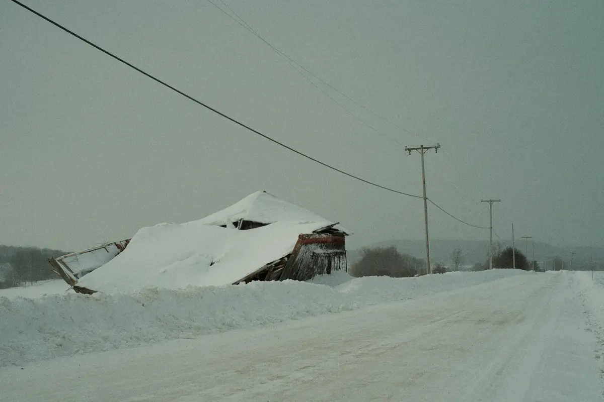 A dilapidated wooden structure, possibly a shed or small barn, is almost entirely buried under a thick blanket of snow, with…