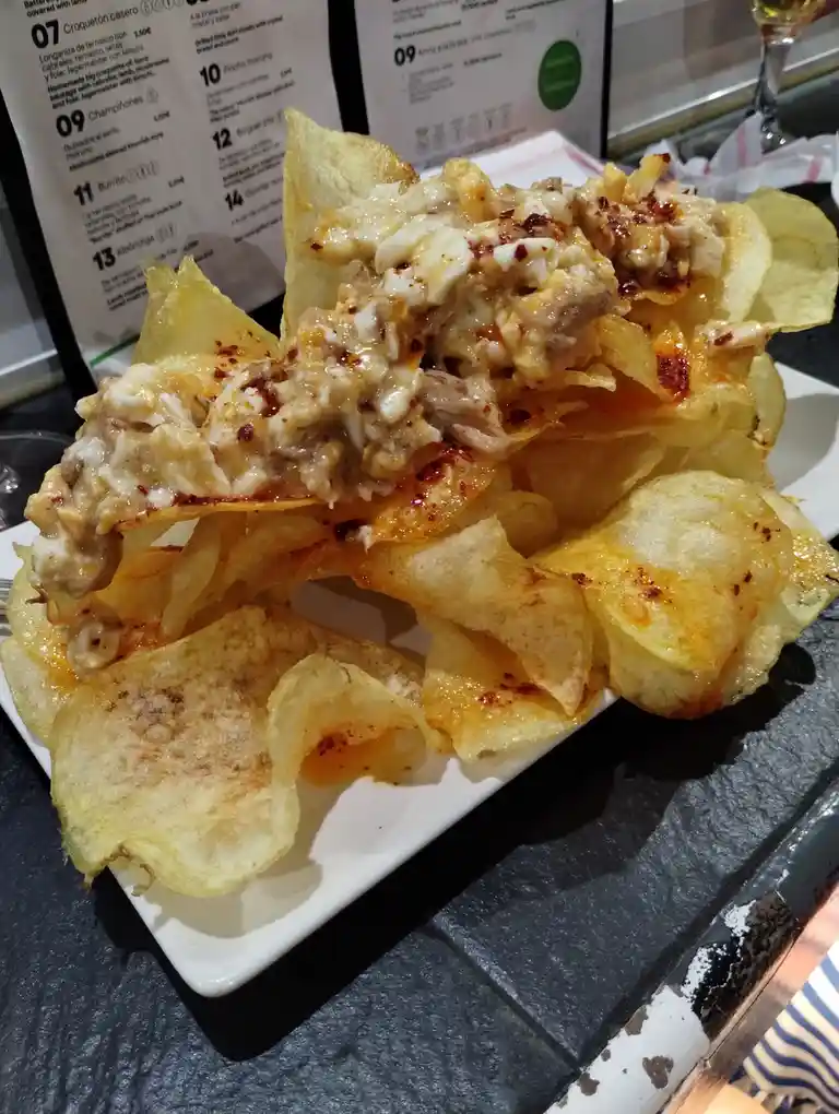A close-up photo shows a plate of loaded potato chips topped with melted cheese, meat, and red seasoning, sitting on a white…