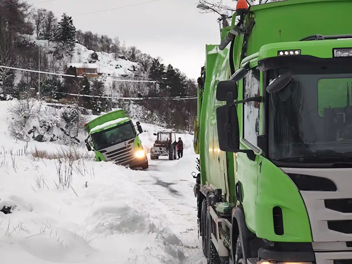 A snowy rural road scene shows two bright green Scania trucks. One truck, labeled '2', is stuck sideways in a deep snowbank…