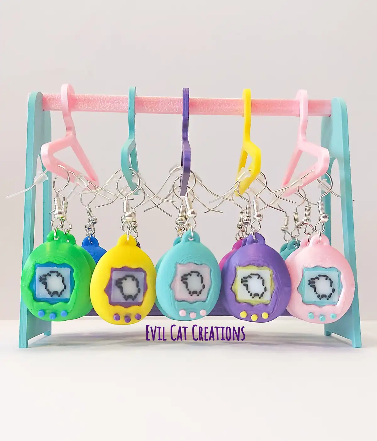 A row of colorful, 3D-printed Tamagotchi-shaped earrings hang from a pink foam rack, each displaying a pixelated 'O' on its…