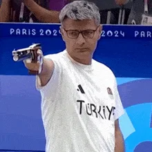 A man in a white 'TURKIYE' t-shirt and glasses is shown in six frames, holding a pistol and aiming it forward with a serious…