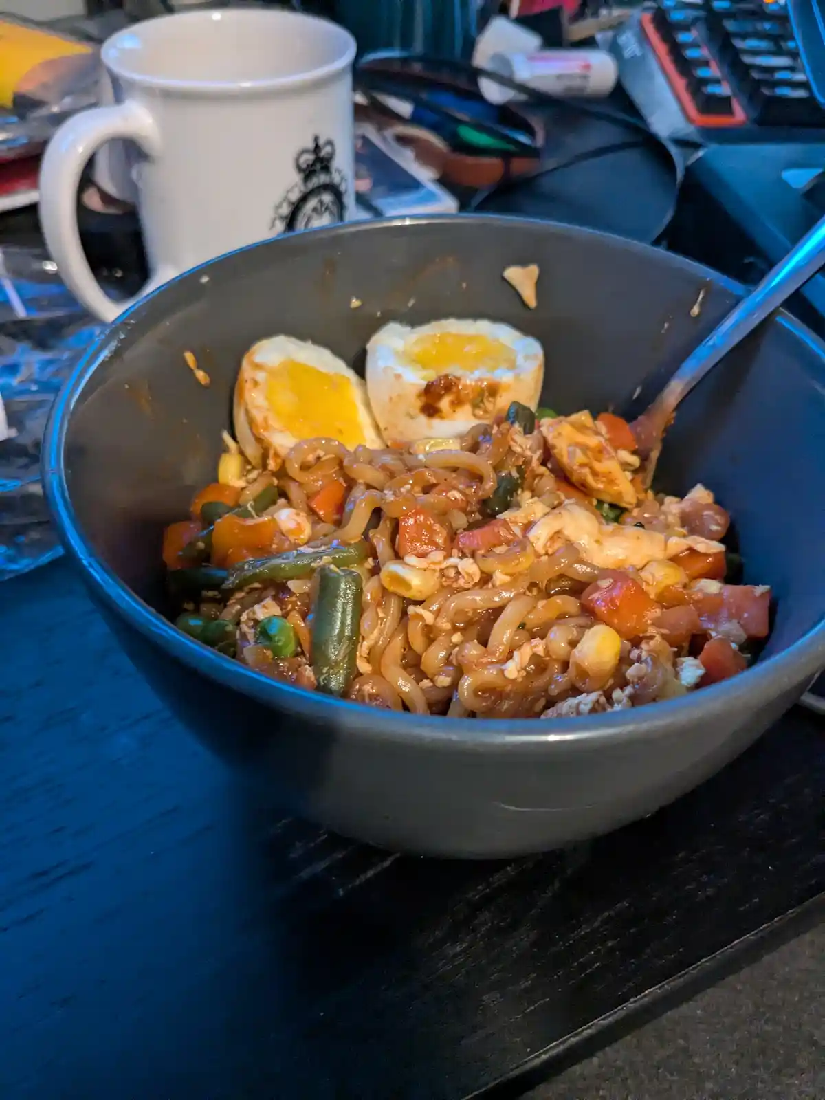 A close-up photo shows a dark bowl filled with stir-fried noodles, mixed vegetables like green beans and corn, and two…