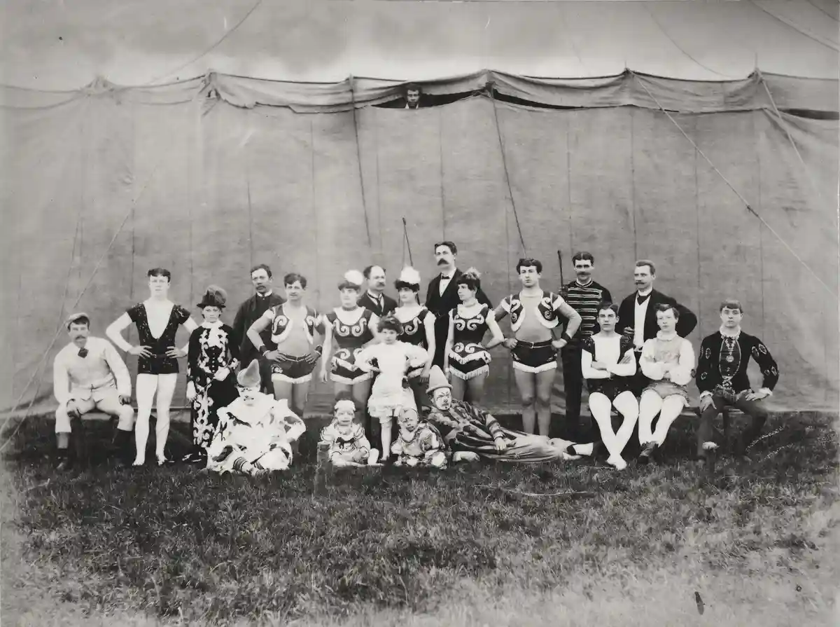 A vintage black-and-white photo shows a group of early 20th-century circus performers posing in front of a large tent. They…