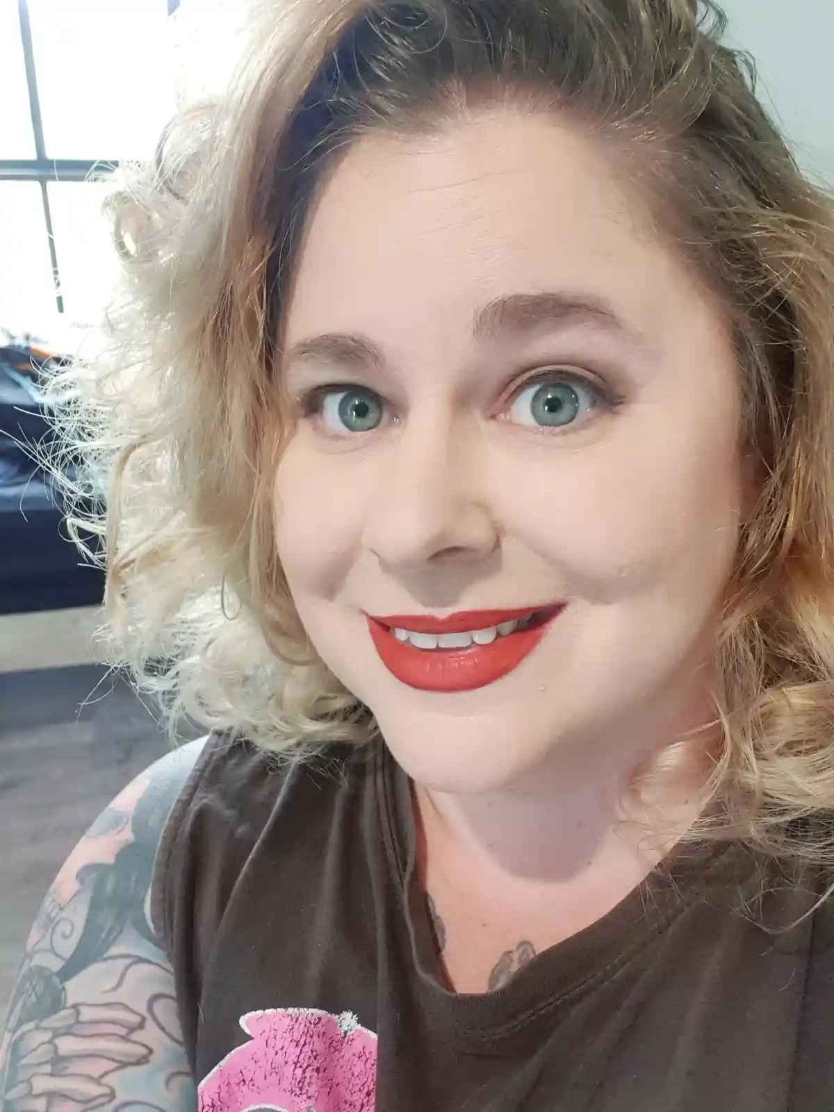 A selfie of a woman with blonde curly hair, green eyes, and bright red lipstick, smiling at the camera. She wears a brown…
