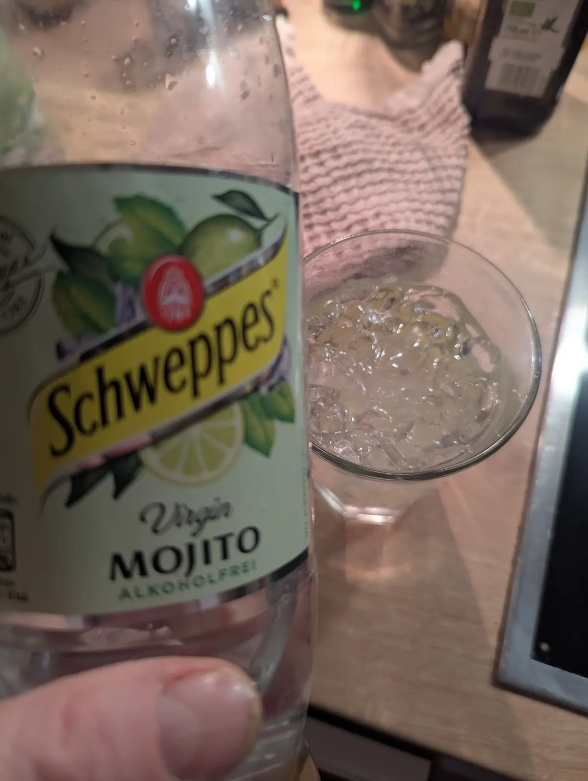 A close-up photo shows a hand holding a Schweppes bottle labeled 'Virgin Mojito Alkoholfrei' next to a glass filled with…