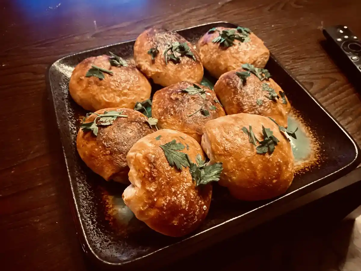 A dark ceramic plate holds eight golden-brown, round bread rolls topped with fresh green parsley, sitting on a wooden table…