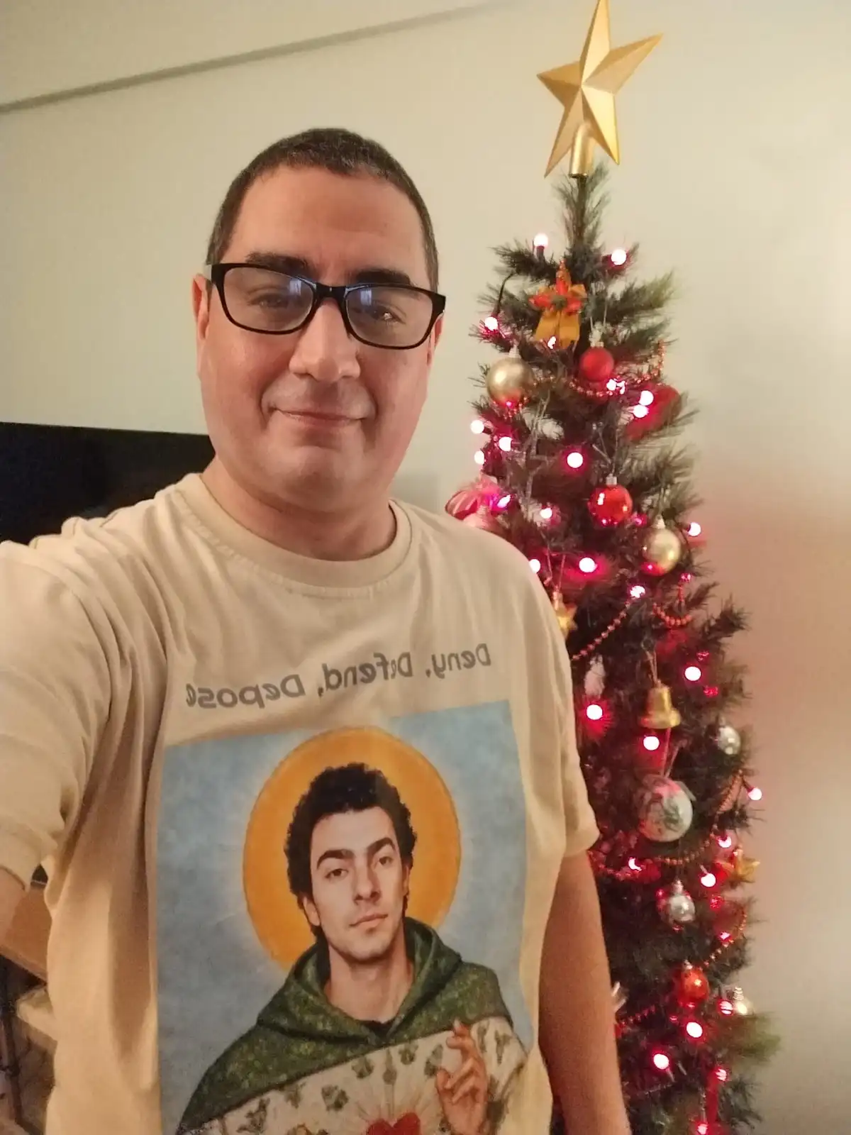 A man wearing glasses and a beige t-shirt with a religious-style portrait of a man and the text 'Deny. Defend. Depose.'…