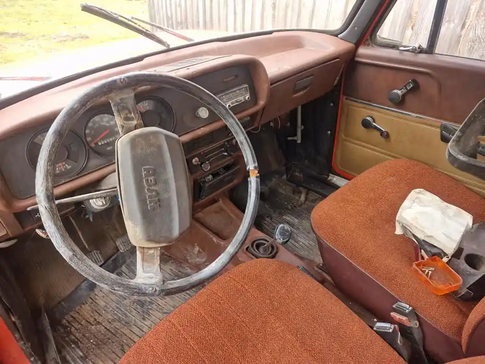 The image shows the interior of a vintage, rusty, and worn-out car with a brown dashboard, faded orange seats, and a…