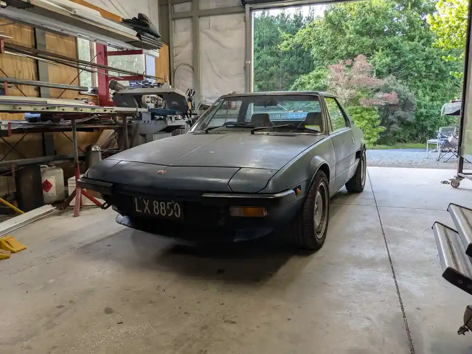 A dusty, dark gray Fiat X1/9 sports car is parked inside a cluttered garage with tools and equipment visible. The car's…