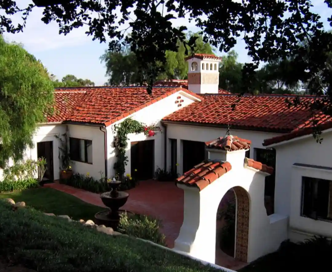 a picturesque white stucco house with red tile roofs, arched doorways, and a central courtyard fountain, surrounded by lush…
