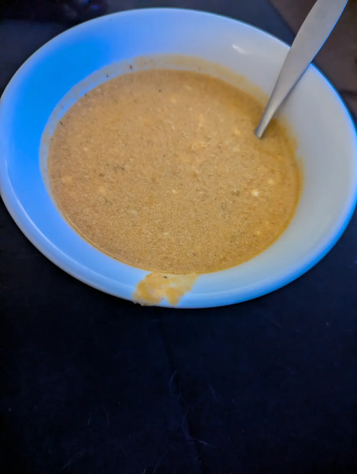 A close-up photo shows a white bowl filled with a thick, orange-brown soup, with a metal spoon resting inside. A small…
