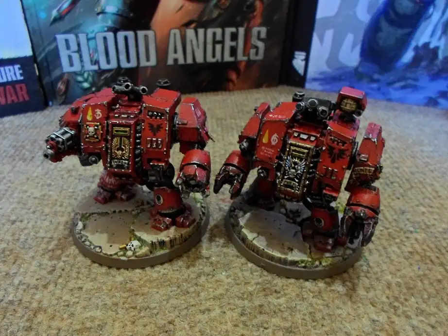 Two red-painted, heavily armored, humanoid war machines stand side-by-side on circular bases, with a 'BLOOD ANGELS' box…