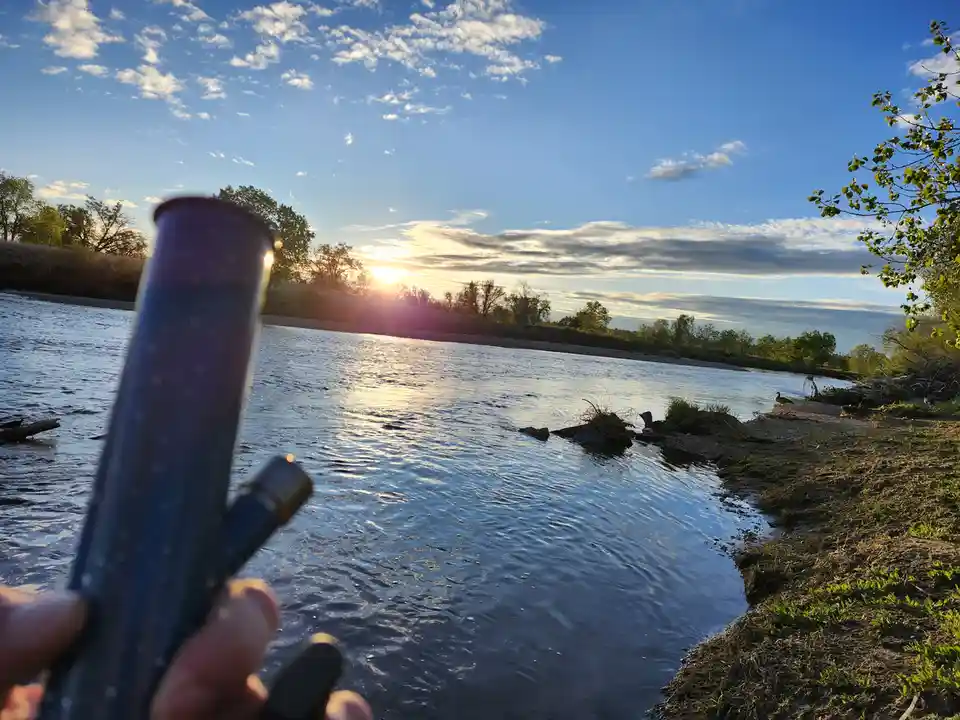 A hand holds a lit vape pen in the foreground, with a serene river and sunset visible behind it. The overlaid text reads…