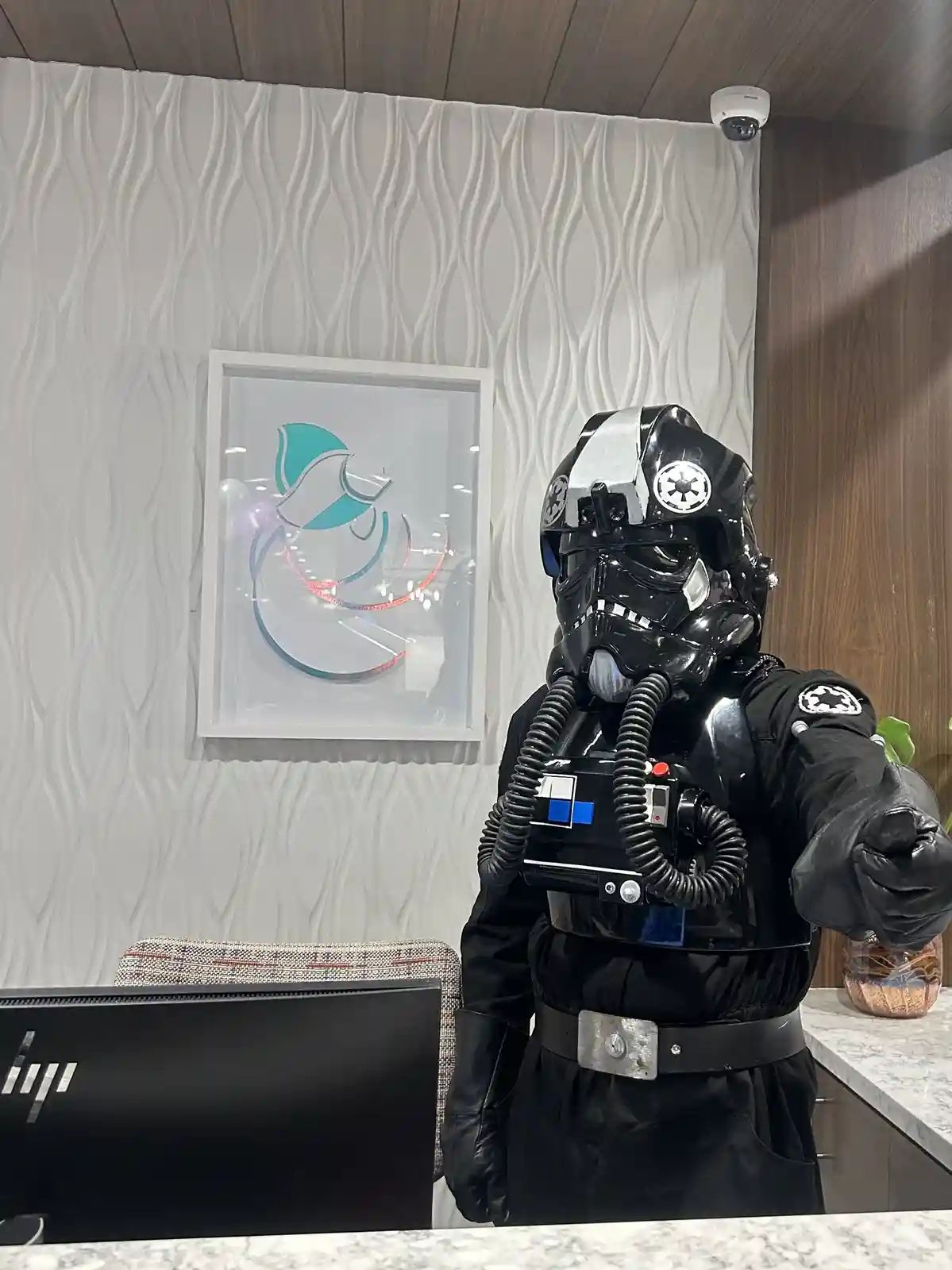 A person in a black stormtrooper costume with a breathing apparatus stands behind a reception desk in a modern office…
