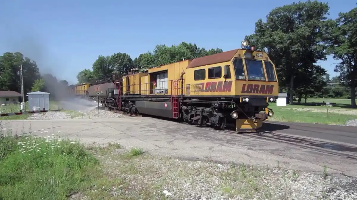 A yellow LORAM railcar, likely a track maintenance or railcar mover, is shown on a track with water spraying from its side…
