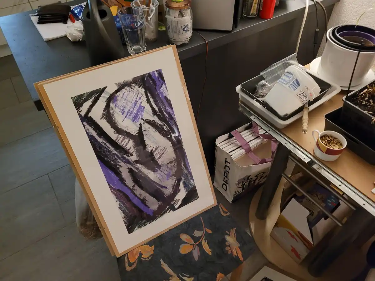 A framed abstract drawing with purple and black strokes leans against a chair in a cluttered room with a kitchen counter, a…