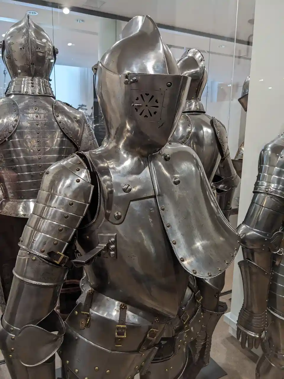 A photo of several suits of medieval armor displayed in a museum, with the central suit facing away from the camera. The…