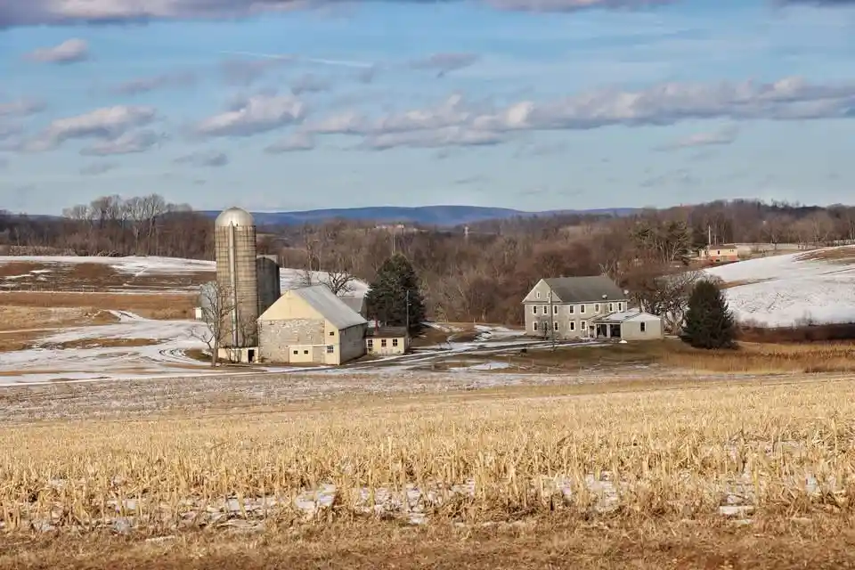 A rural farm scene with a silo, barns, and houses surrounded by snowy fields and bare trees under a partly cloudy sky…