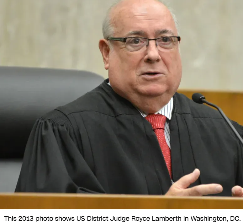 A man in a black judicial robe and red tie sits at a wooden bench with a microphone, gesturing with his right hand while…