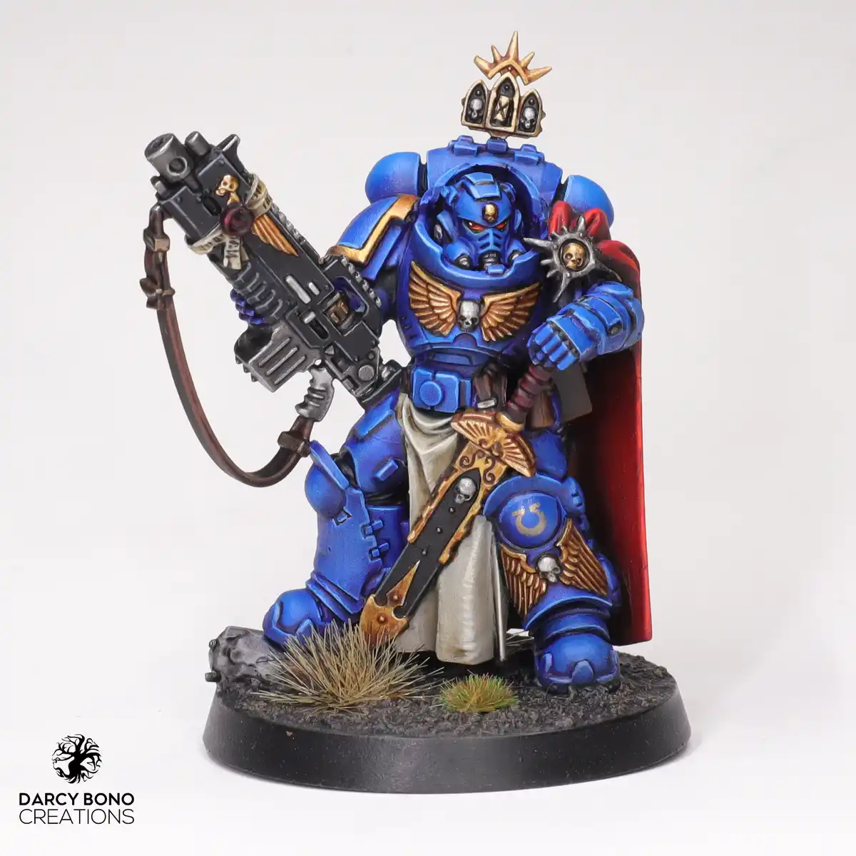 A highly detailed, blue-painted miniature of a Space Marine from Warhammer 40,000, kneeling on a rocky base with sparse…