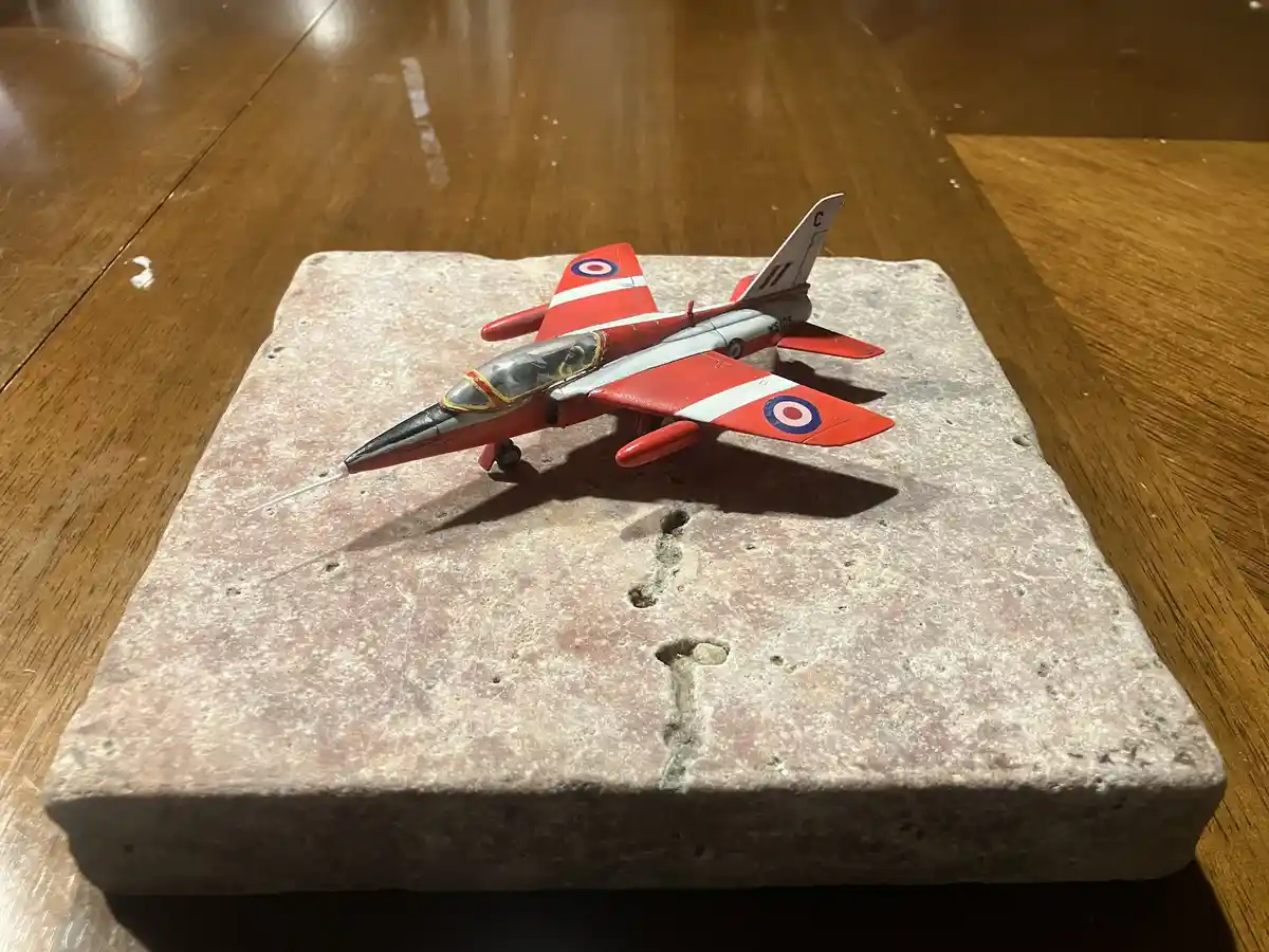 A red and white model airplane with British roundel markings sits on a rough, weathered stone block, which has a visible…