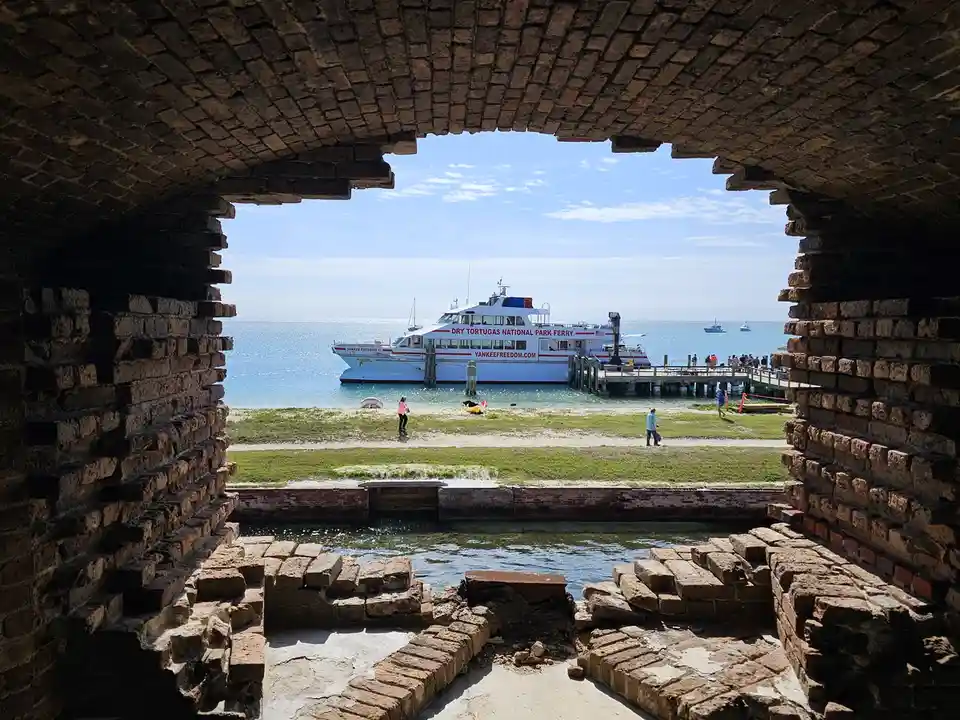 A view from inside a weathered brick structure looking out onto a sunny beach where a white ferry labeled 'DRY TORTUGAS…