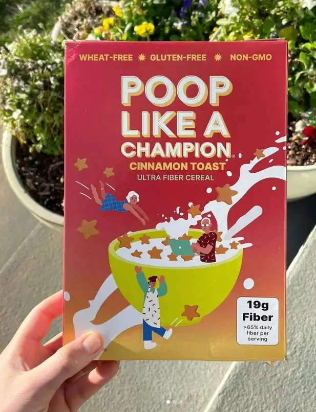 A hand holds a red cereal box labeled 'POOP LIKE A CHAMPION. CINNAMON TOAST ULTRA FIBER CEREAL' with claims of being…