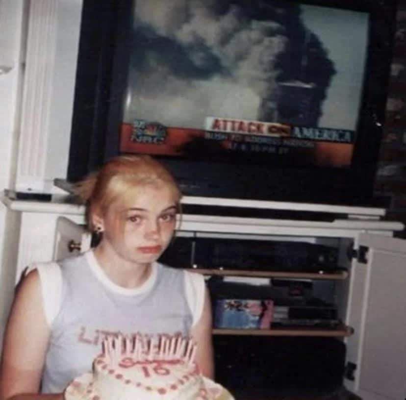 A young person with blonde hair sits in front of a TV showing news footage of the 9/11 attacks, holding a birthday cake with…