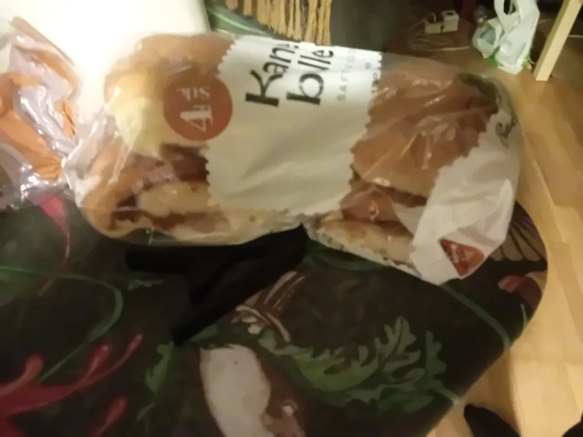 A blurry photo shows a bag of 'Yamie' brand sandwich rolls sitting on a patterned cushion, with a black sock or piece of…