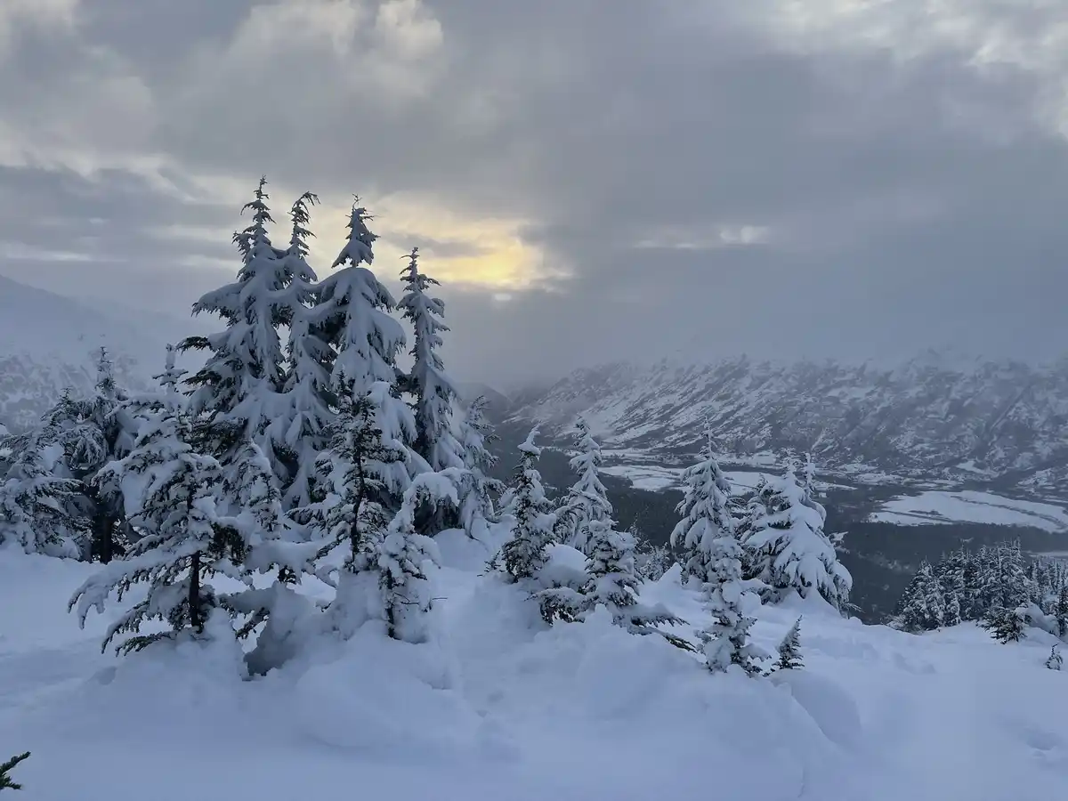 A serene, snow-covered mountain landscape with evergreen trees heavily laden with snow, under a cloudy sky with a hint of…