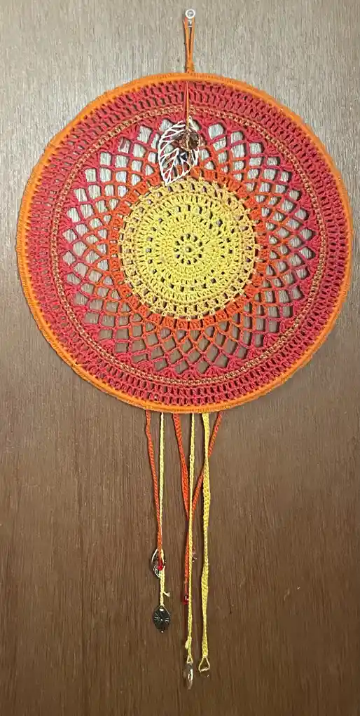 A hand-crocheted dreamcatcher with a gradient of orange, red, and yellow yarn, hanging on a wooden surface. It features a…
