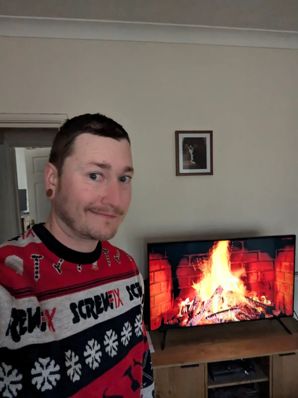 A man wearing a red, black, and white Christmas sweater with 'Screwfix' printed on it smiles at the camera while standing in…
