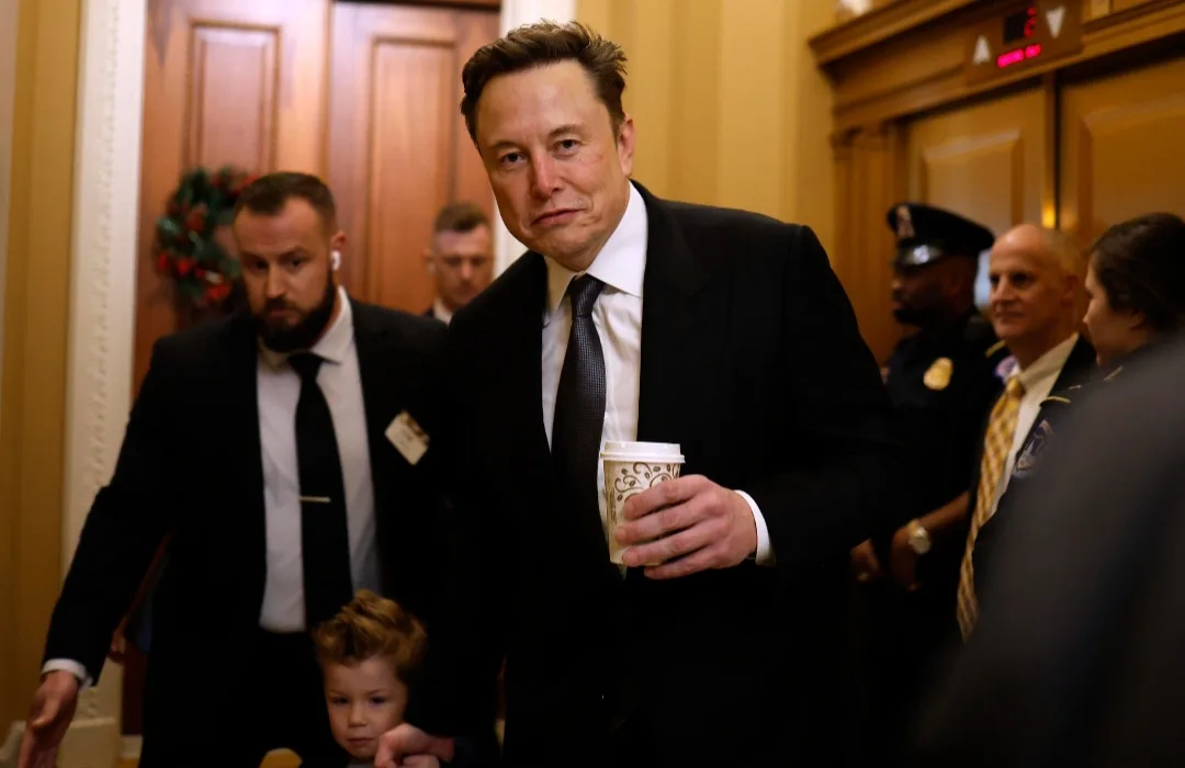 elon musk walks down a hallway in a suit holding a coffee cup, looking tired or annoyed, with security and other people…